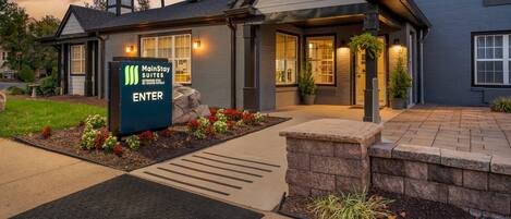 A MainStay Suites hotel entrance with a sign, a covered walkway, and a landscaped area with plants.