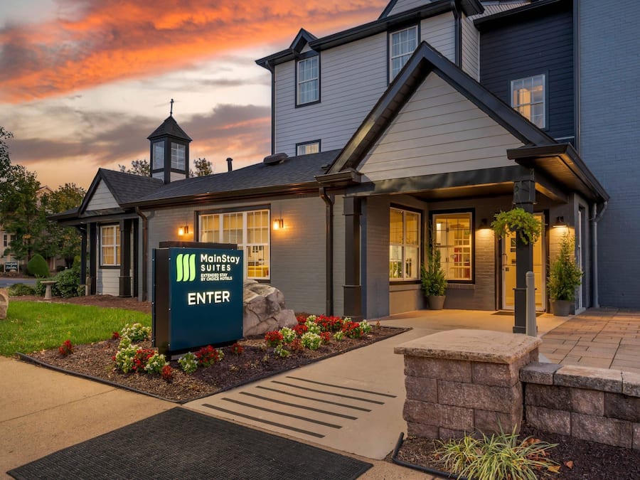 A MainStay Suites hotel entrance with a sign, a covered walkway, and a landscaped area with plants.