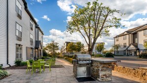 Une terrasse avec des sièges extérieurs et un barbecue, entourée d’immeubles résidentiels et d’un arbre.
