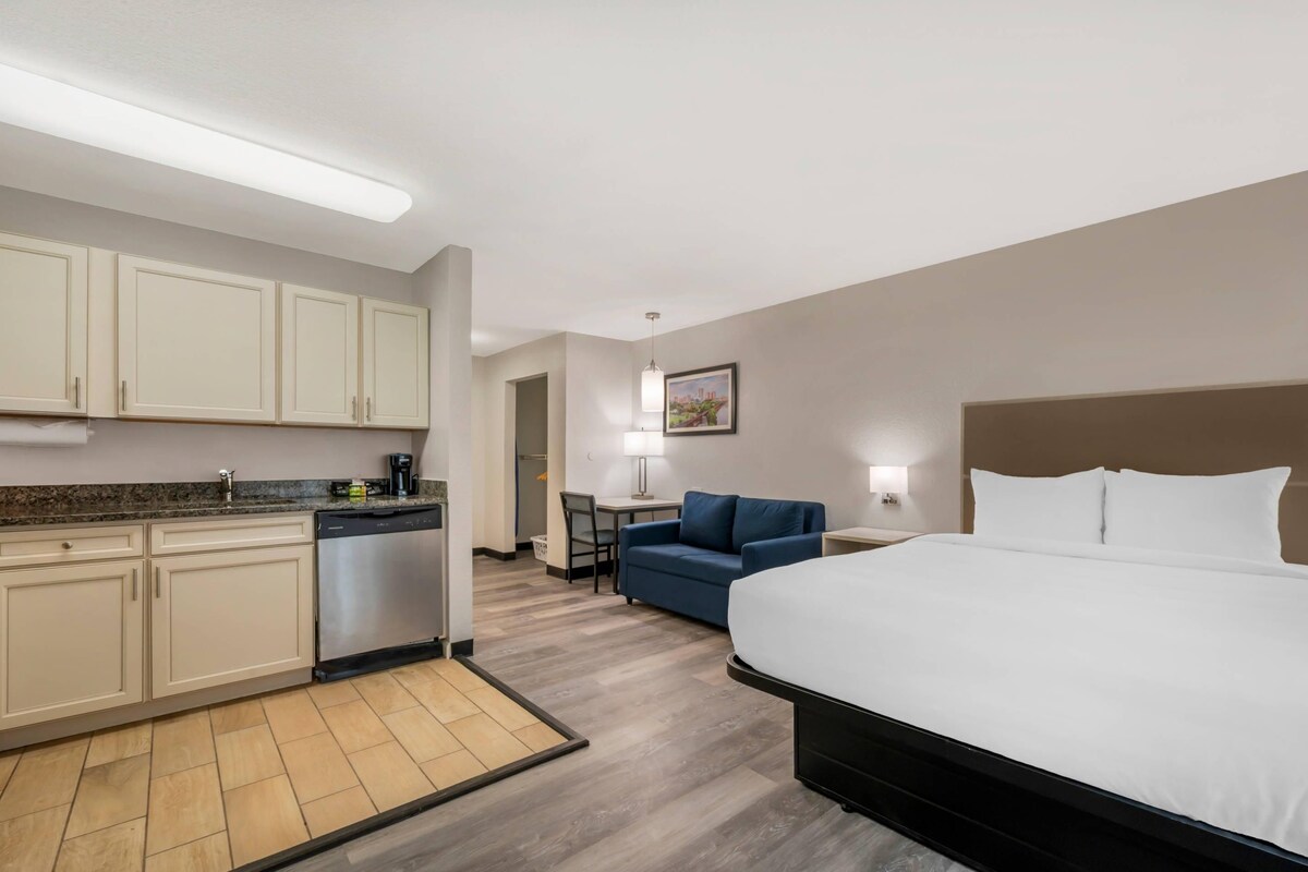 A hotel room with a large bed, a kitchenette with a dishwasher, a blue sofa, and a small table with a lamp.