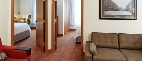 Una habitación de hotel con una cama, un sillón rojo, un sofá marrón, un cuadro en la pared y una alfombra estampada.