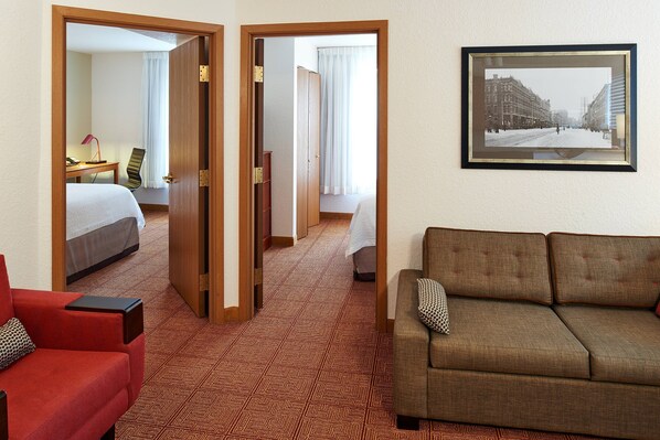 Una habitación de hotel con una cama, un sillón rojo, un sofá marrón, un cuadro en la pared y una alfombra estampada.