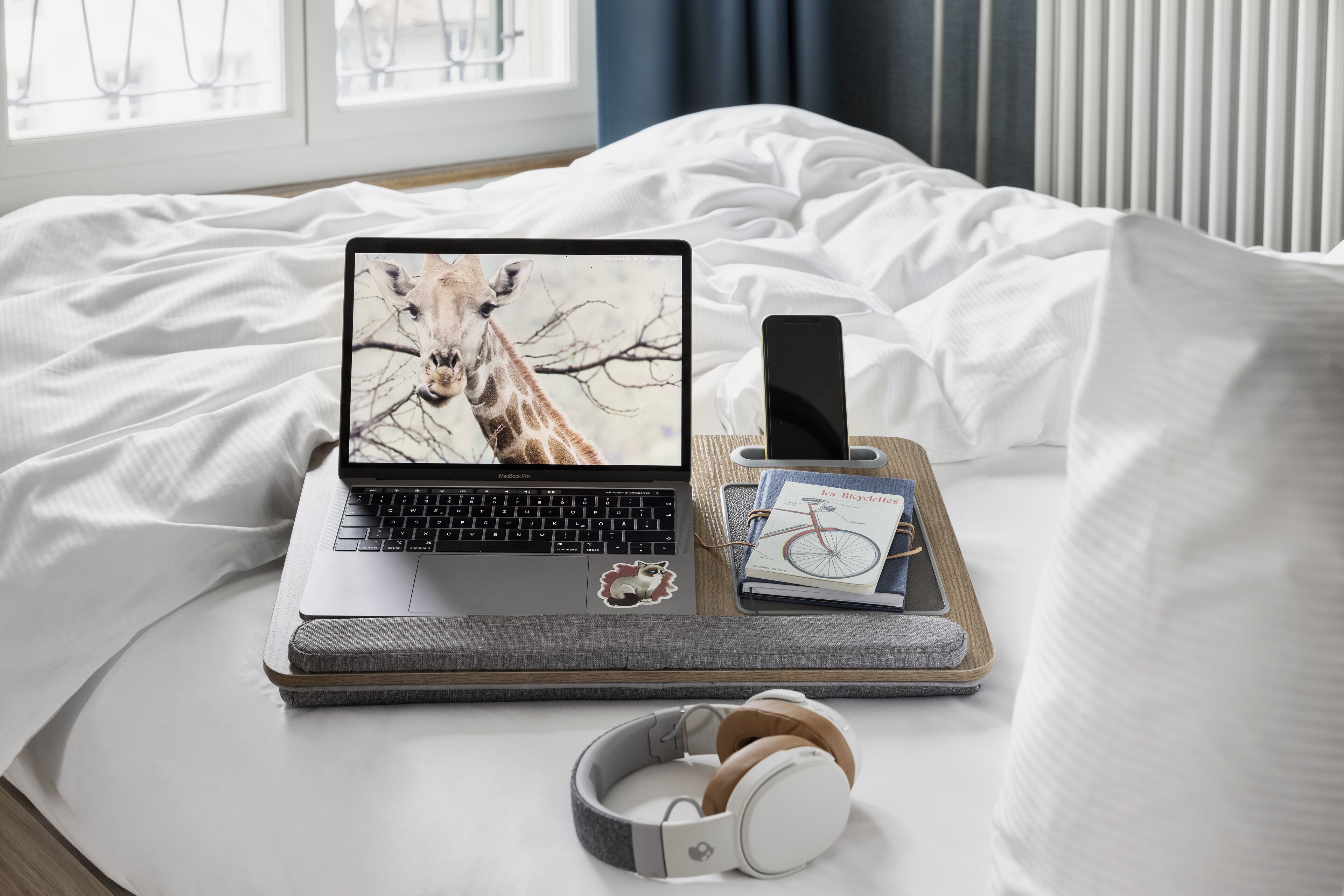 A laptop on a bed with a giraffe sticker, a smartphone, a book, and headphones.