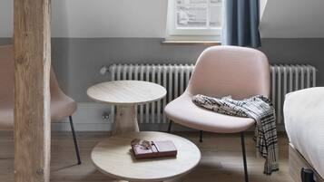 Un dormitorio estilo loft con vigas de madera a la vista, una silla rosa, una mesa redonda y vistas de un paisaje urbano a través de una ventana con cortinas azules.