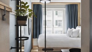 Habitación de hotel con cama, una planta en un estante, una lámpara de pie y vistas a edificios a través de la ventana.