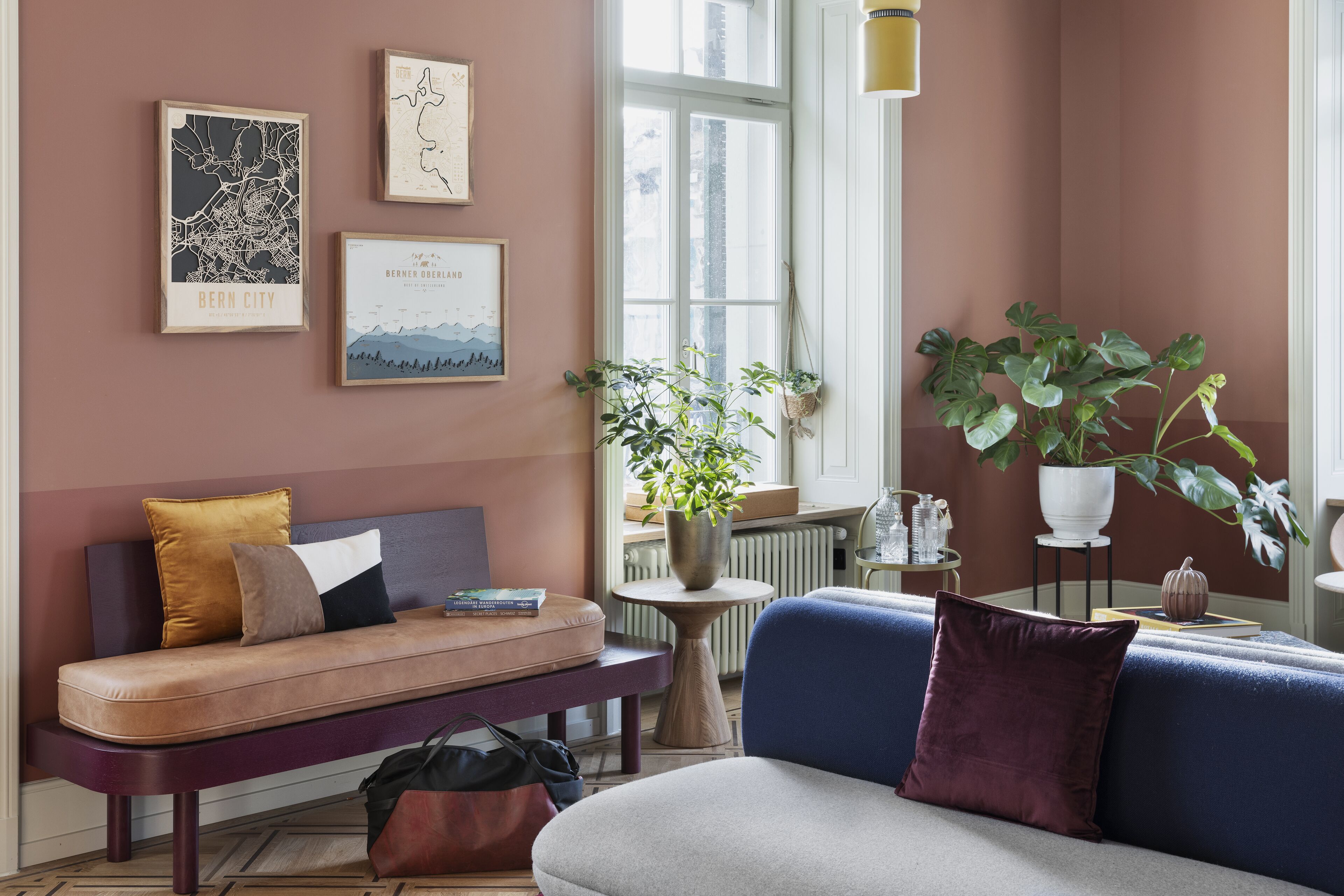 Un salon chaleureux avec un canapé, un banc et une table basse. Des œuvres d’art encadrées ornent le mur, et une plante en pot est posée sur le rebord de la fenêtre.