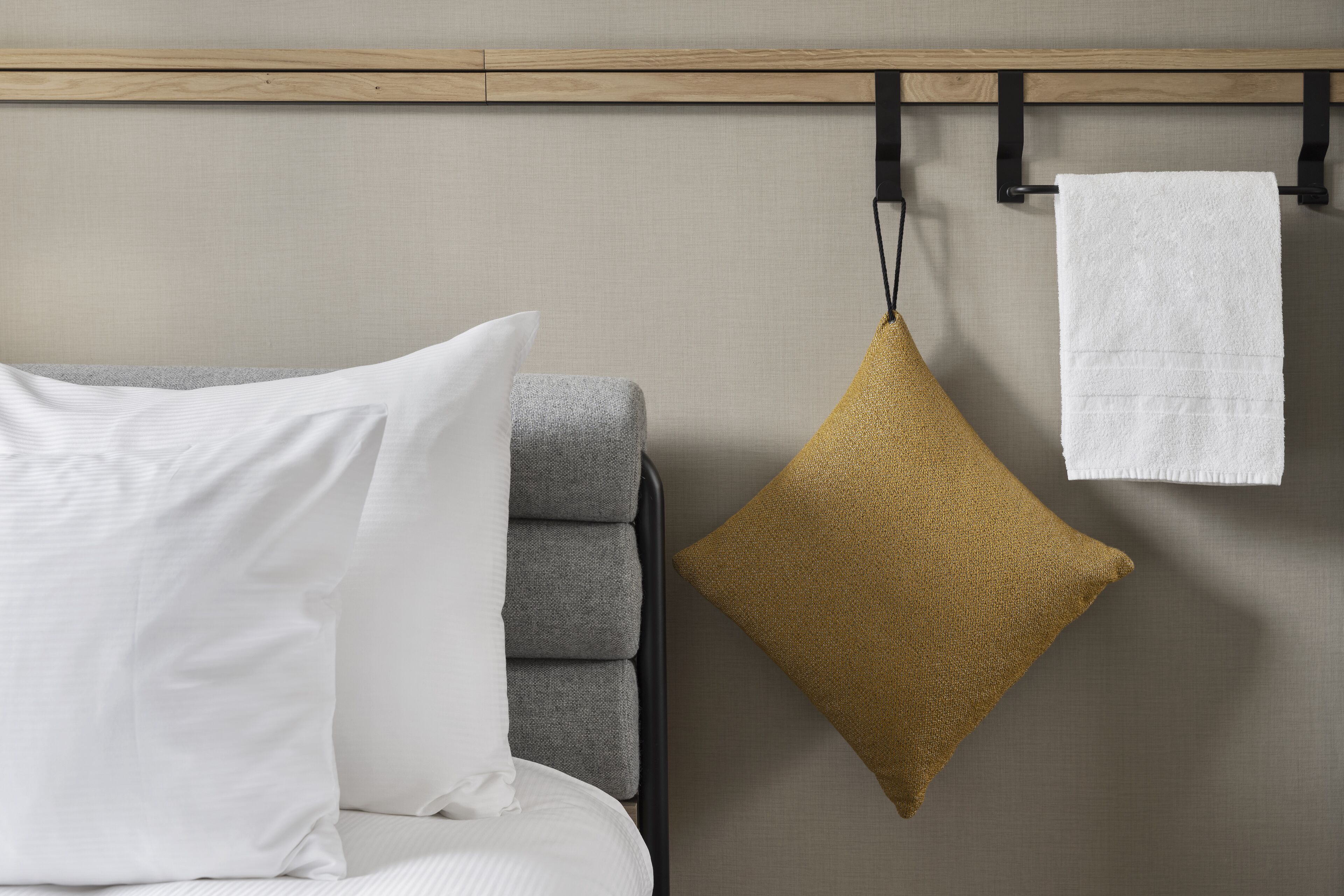 Una cama con ropa de cama blanca y gris, una almohada dorada y una toalla blanca colgada en un perchero negro.