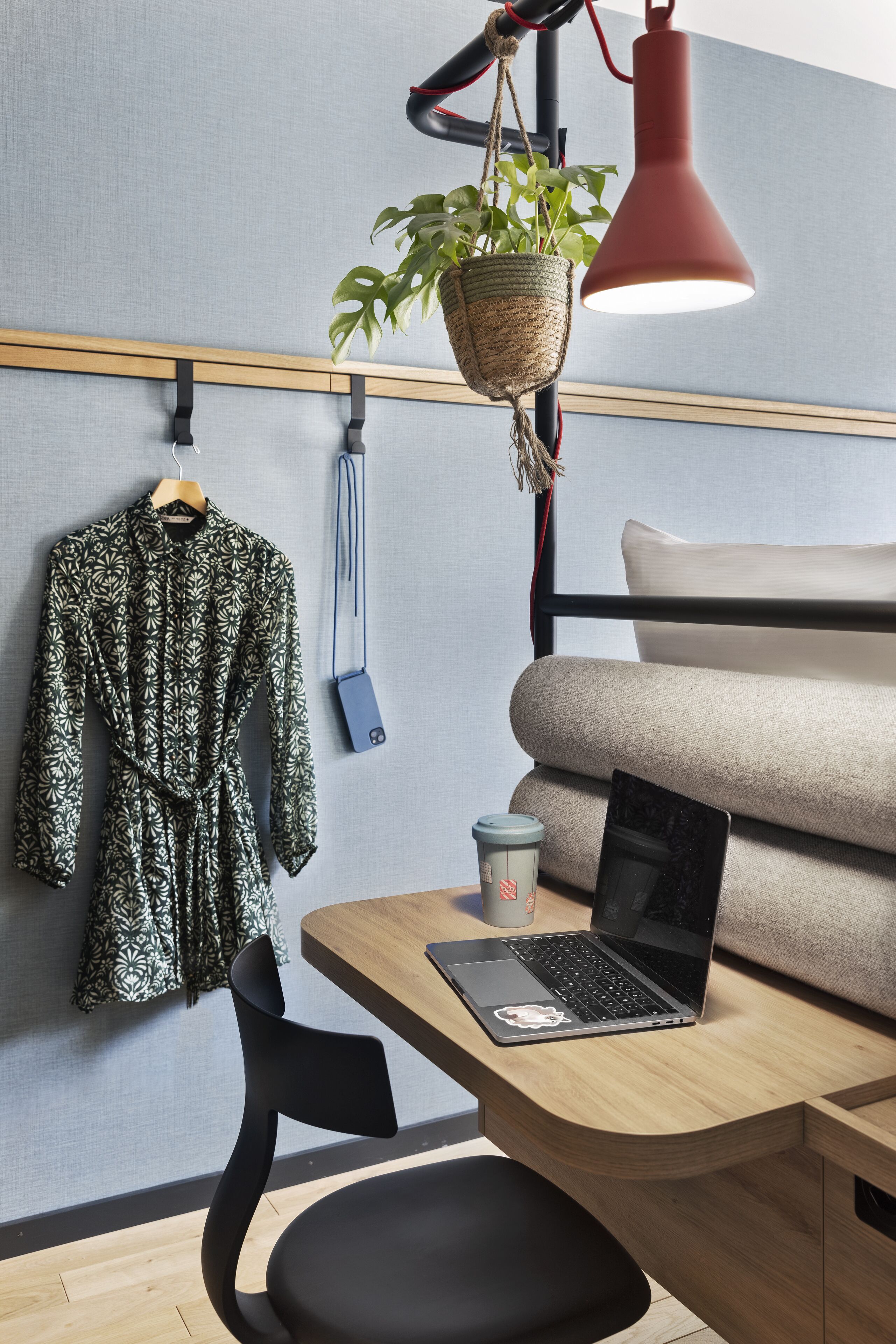 Une chambre moderne avec un bureau en bois, un ordinateur portable, une plante suspendue, un manteau sur un cintre et un canapé.