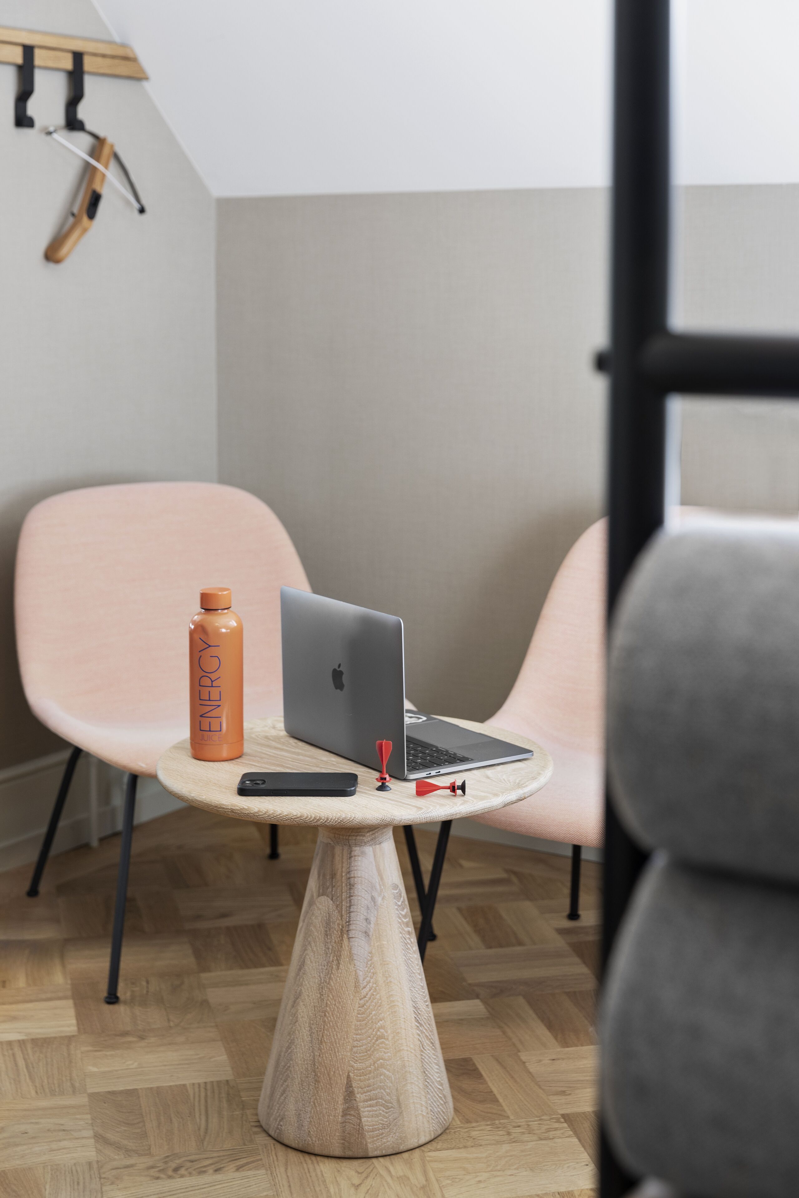 Una habitación moderna con una laptop sobre una mesa redonda de madera, un teléfono inteligente y una botella de agua naranja con la etiqueta ENERGY. Hay dos sillas, una rosa y otra gris, y un suelo de madera.