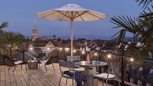A rooftop terrace with outdoor seating, string lights, and a view of a cityscape including a church tower. - Stay KooooK Bern City – Online Check In (Bern)