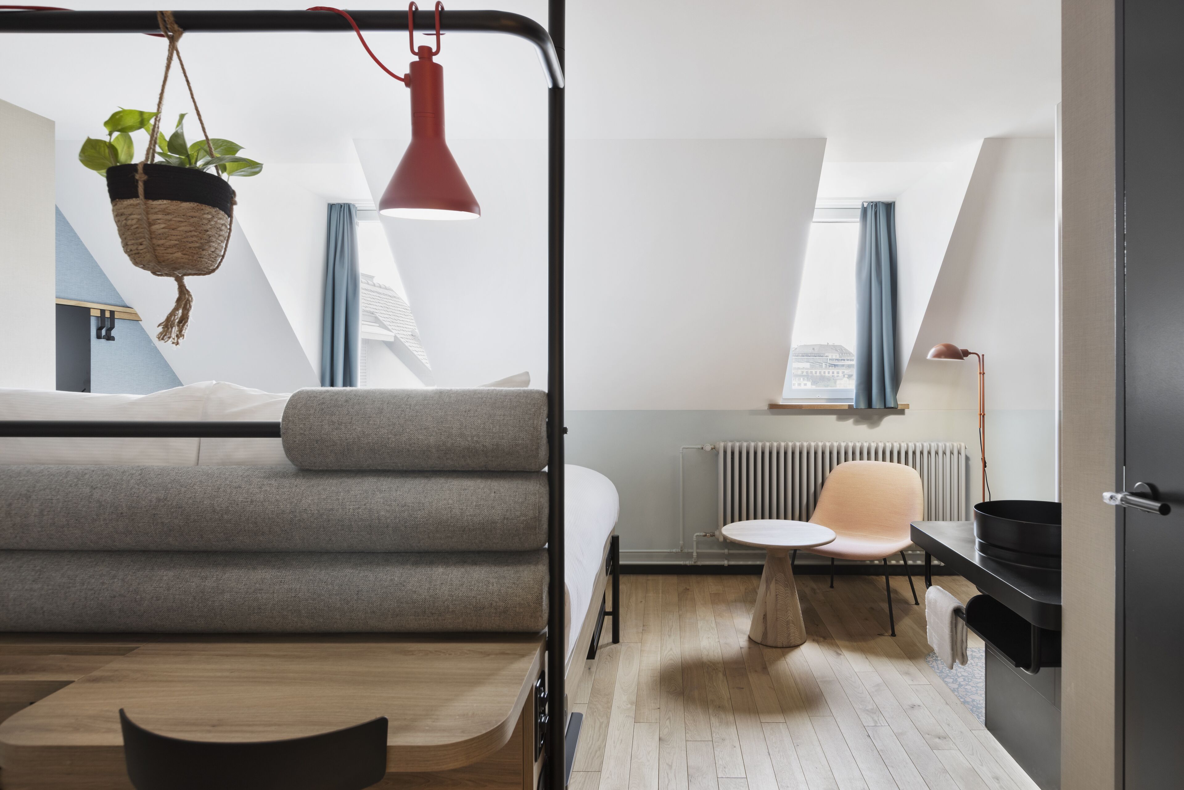 Habitación moderna y compacta con cama, una mesita, una silla, una planta colgante y una lámpara colgante roja.