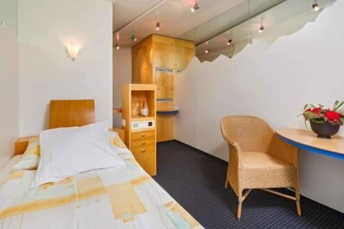 A hotel room with a bed, a wooden wardrobe, a wicker chair, and a small table with a plant.