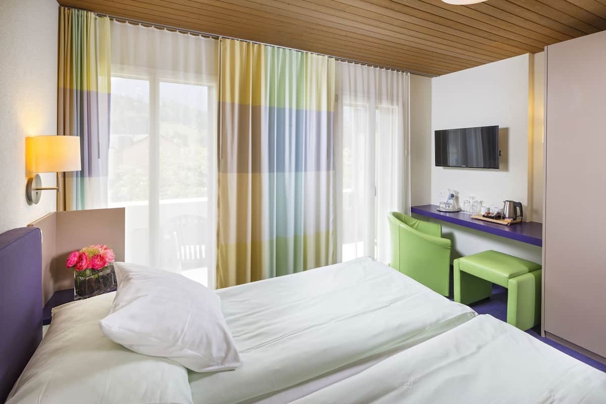 A hotel room with a bed, a desk with a flat-screen TV, a chair, and a small table with a kettle and cups.