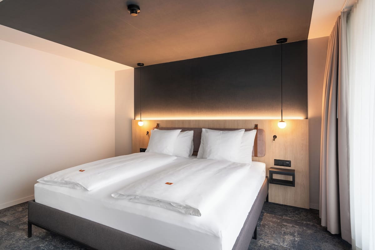 A modern hotel room with a large bed, two bedside tables, and a headboard with integrated lighting.