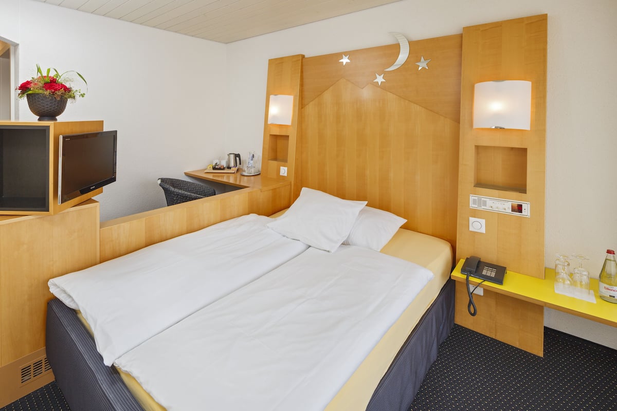 A hotel room with a bed, a desk with a phone, a TV, and a small table with a lamp and a bottle.