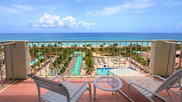 A balcony with white metal chairs and a small round table, overlooking a pool and beach area with palm trees and a turquoise pool. - Sea View Hotel, Bal Harbour, On The Ocean (Bal Harbour)