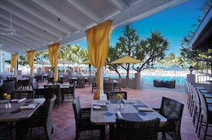 An outdoor dining area with wicker furniture, yellow curtains, and a view of a pool and palm trees.