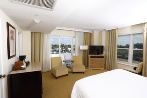 A hotel room with a bed, a desk with a coffee maker, two armchairs, a TV, and a window with curtains.