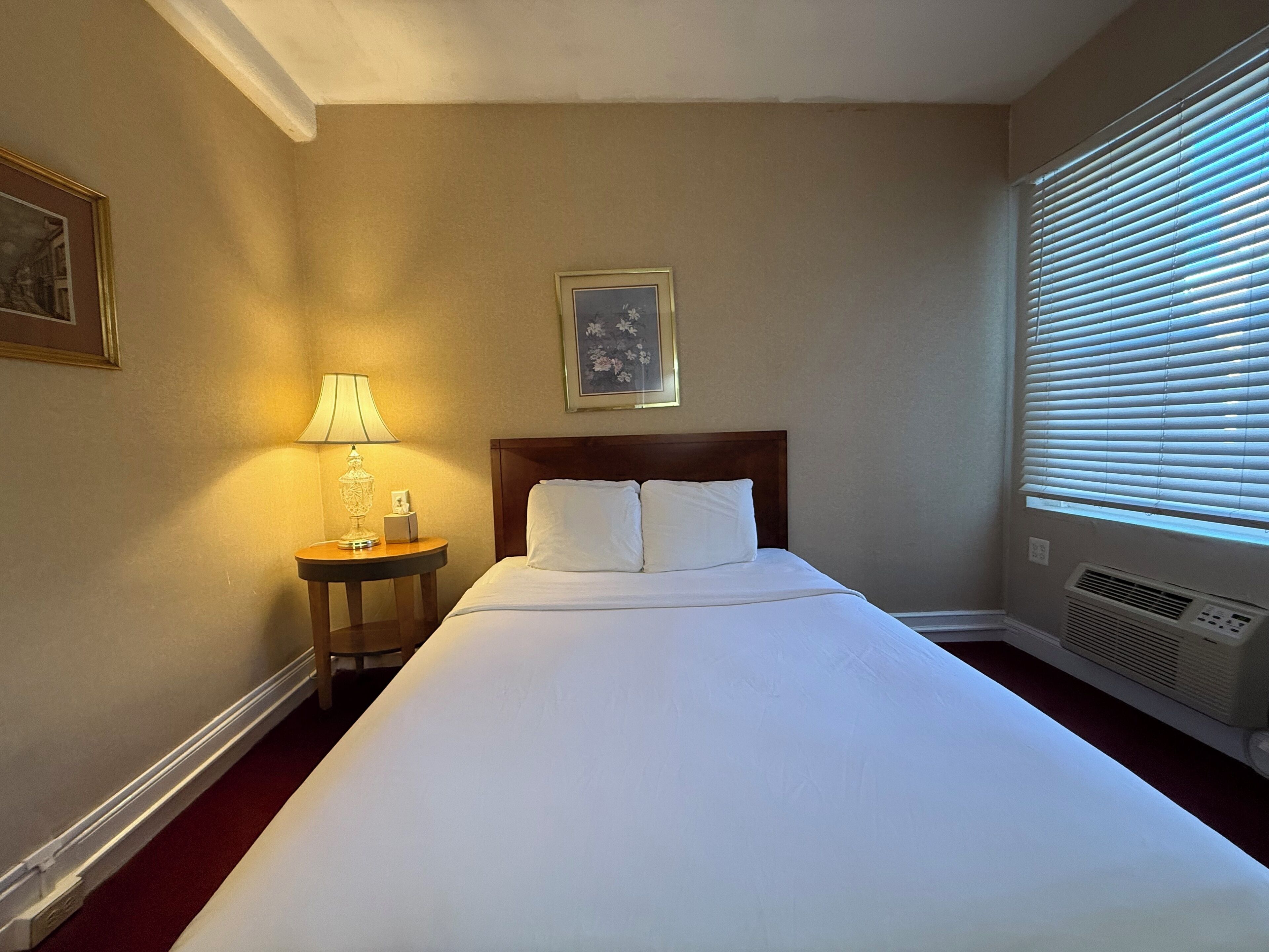 A hotel room with a large bed, a bedside table with a lamp, a framed picture on the wall, and a window with blinds.