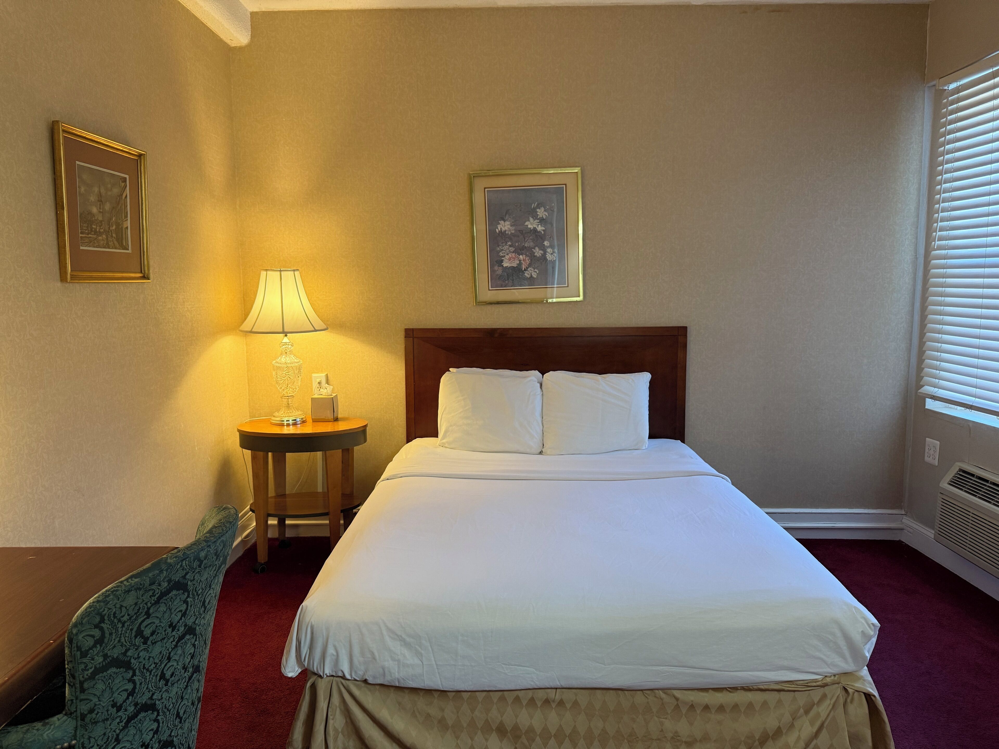 A hotel room with a bed, a nightstand with a lamp, a chair, and two framed pictures on the wall.
