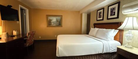 Quarto de hotel com cama, escrivaninha, cadeira, televisão e um quadro na parede.