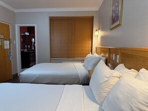 A hotel room with two single beds, a wooden headboard, a window with blinds, a desk with a lamp, and a framed picture on the wall.
