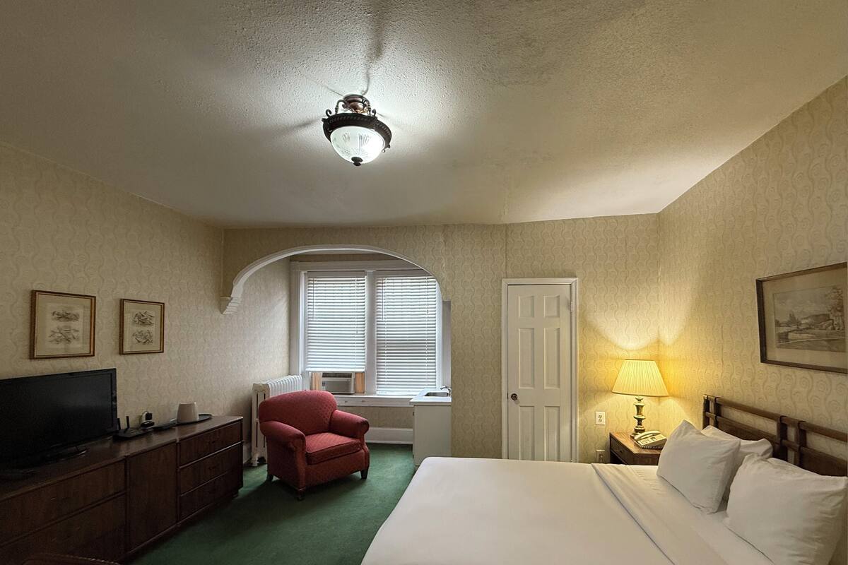 A hotel room with a bed, a desk with a TV, a chair, a lamp, and a window with blinds.