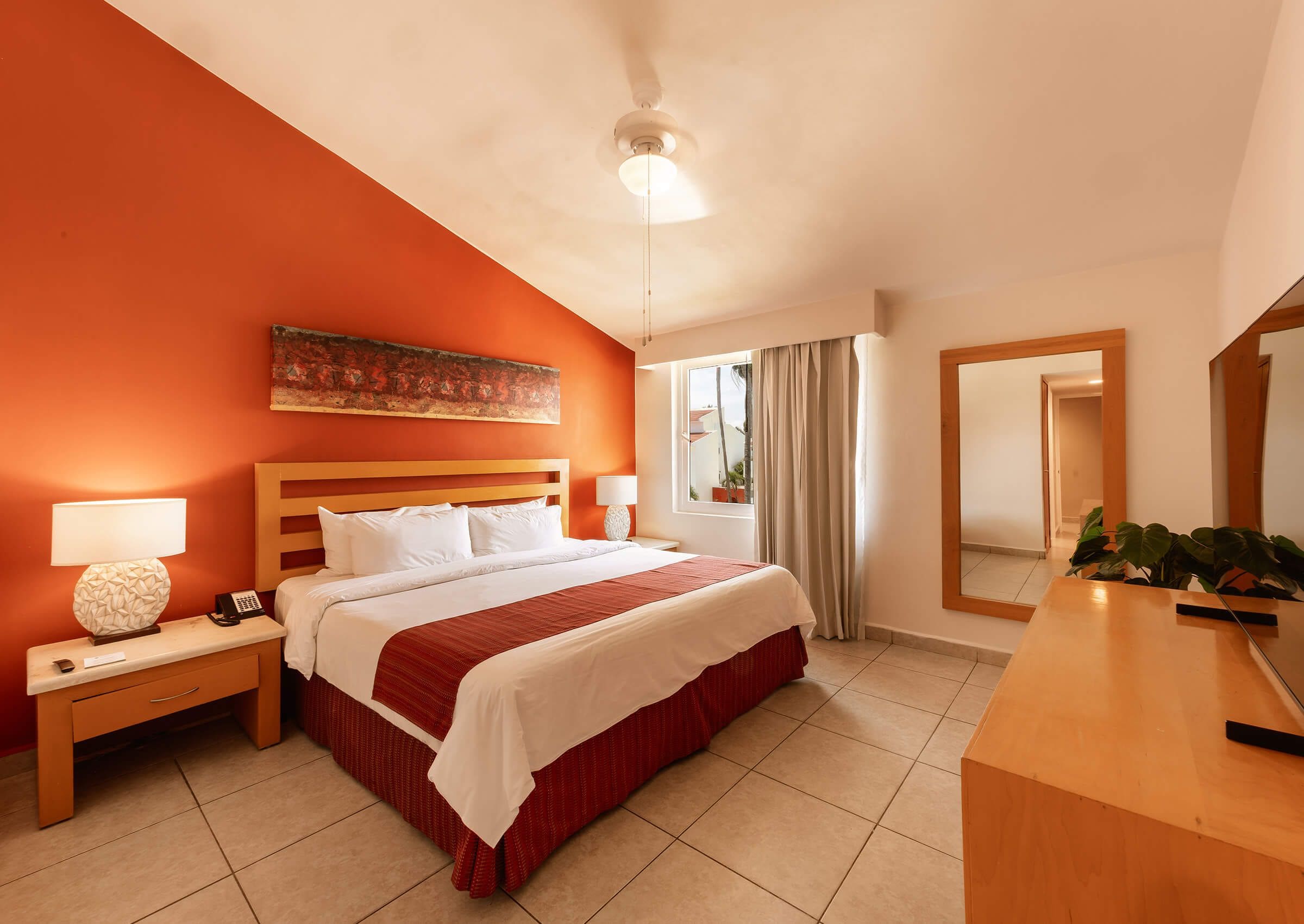 Una habitación de hotel con cama, mesitas de noche, escritorio, espejo y ventana con cortinas.