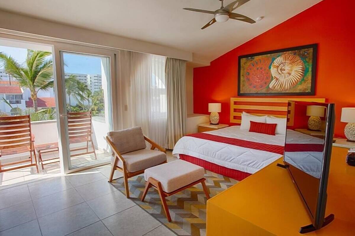A hotel room with a bed, a chair, a desk, a ceiling fan, and a large window with curtains.