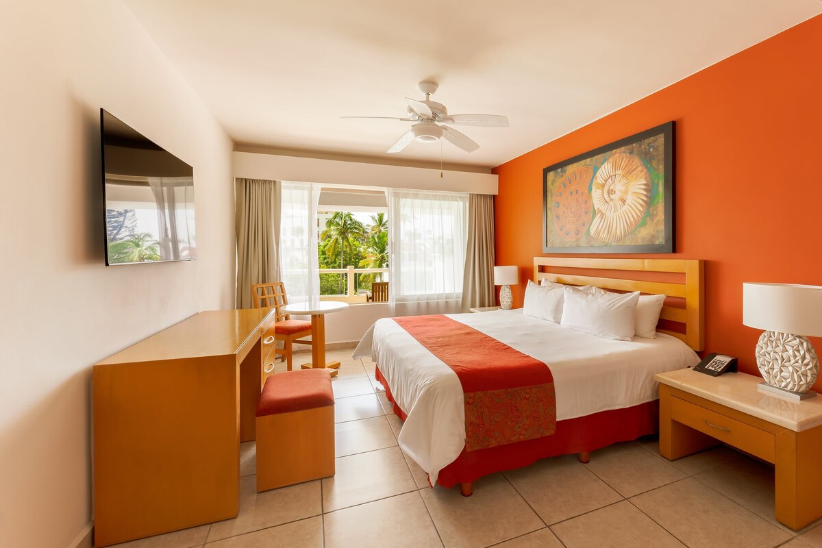 Habitación de hotel con cama, escritorio, silla y vista a palmeras a través del ventanal.