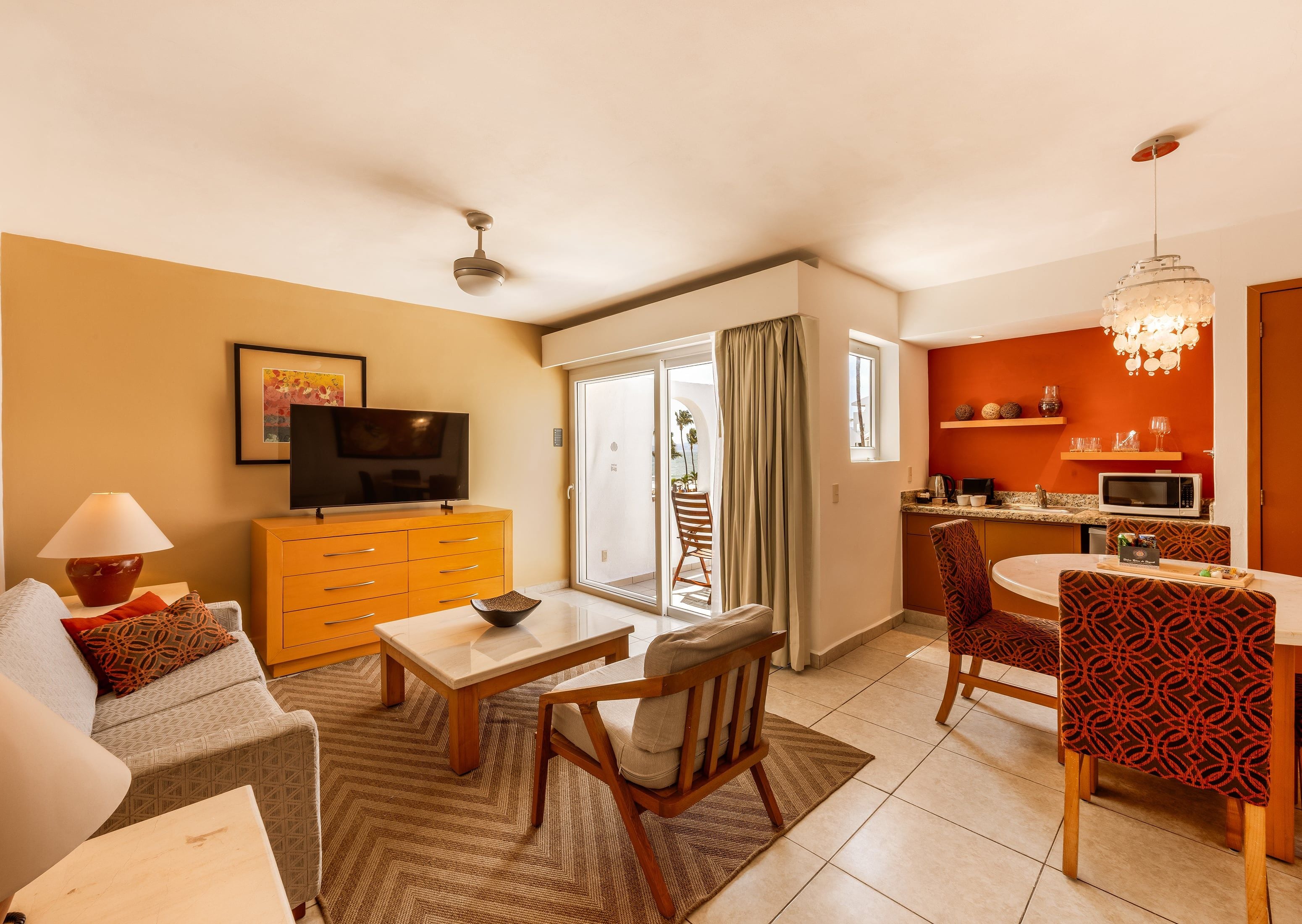 Una sala de estar moderna con sofá, mesa de centro y televisor, un comedor con sillas, una cocina con microondas y un balcón con vistas.