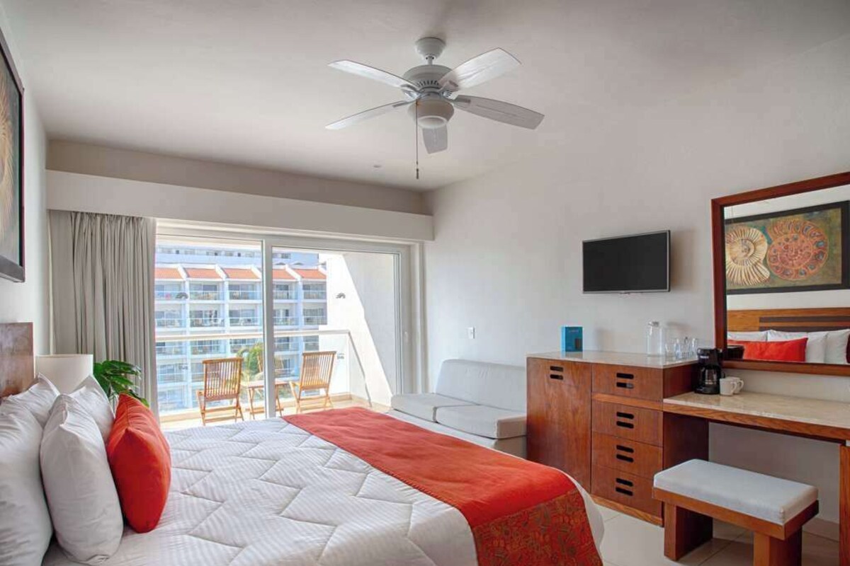 Habitación de hotel con cama, escritorio, silla, televisor y balcón con vista a edificios.