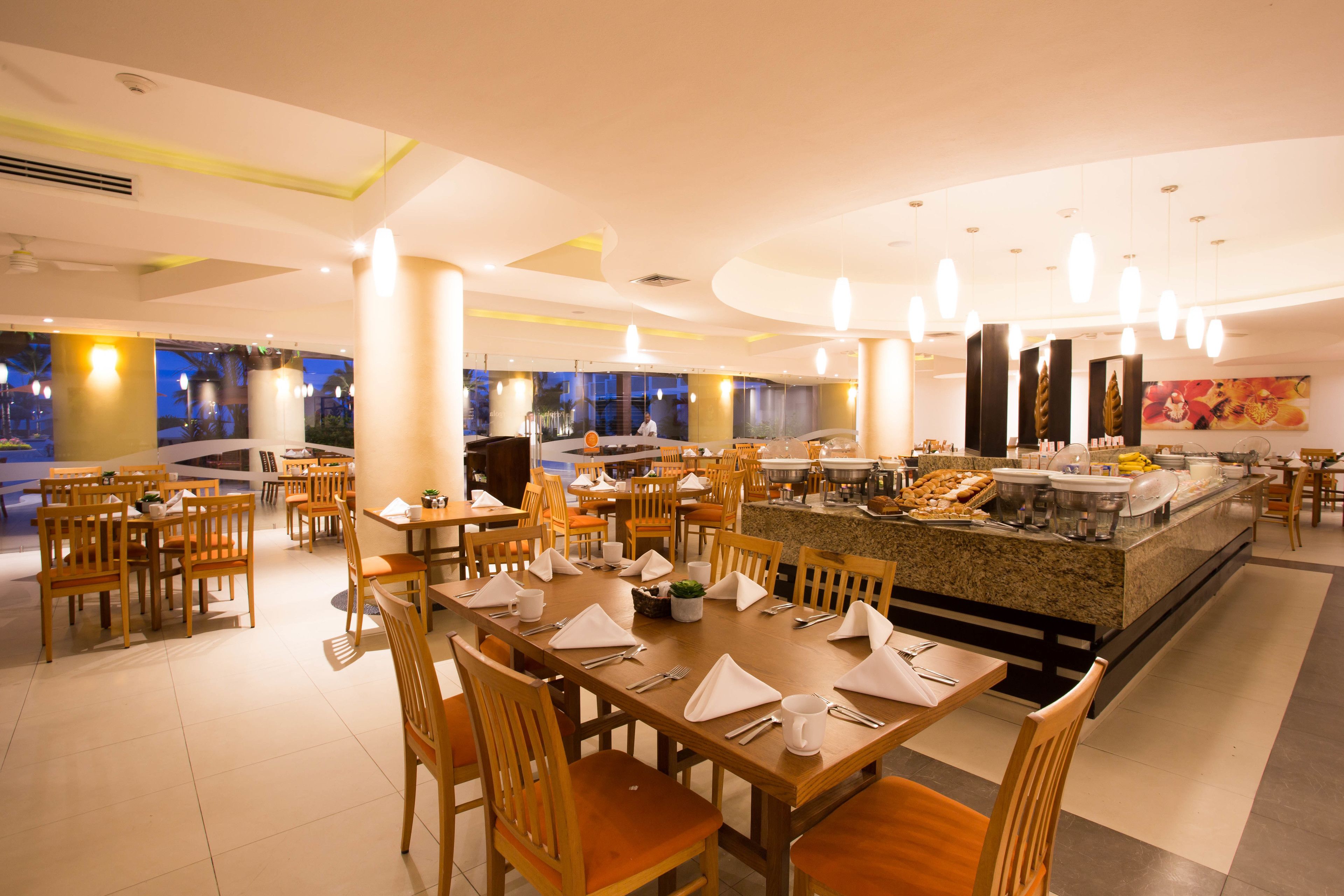 Un restaurant moderne avec des tables et des chaises en bois, un buffet central et une vue sur le paysage urbain illuminé la nuit.