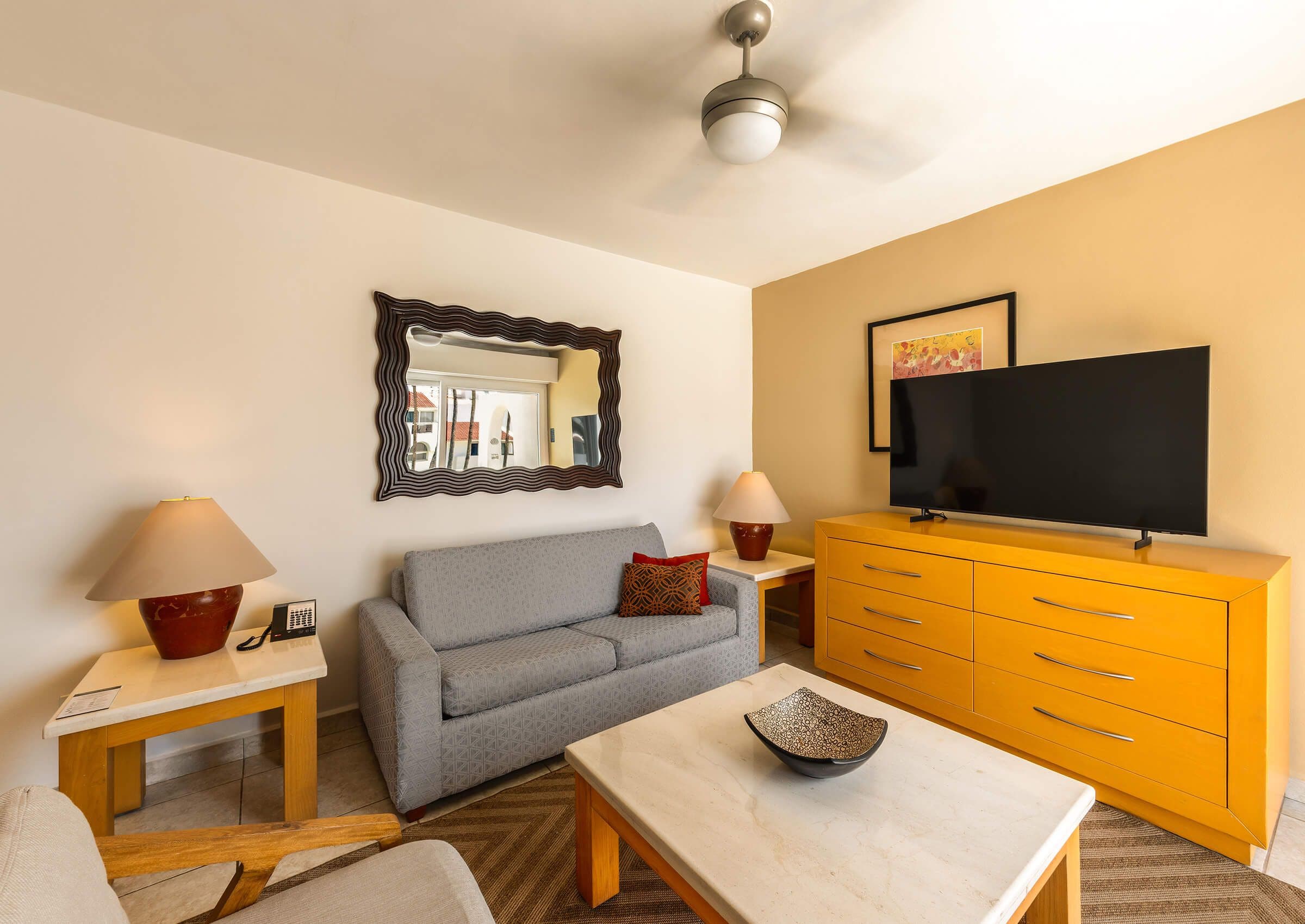 Una sala de estar con un sofá gris, una mesa de centro de madera, un mueble amarillo, un televisor de pantalla plana y un ventilador de techo.
