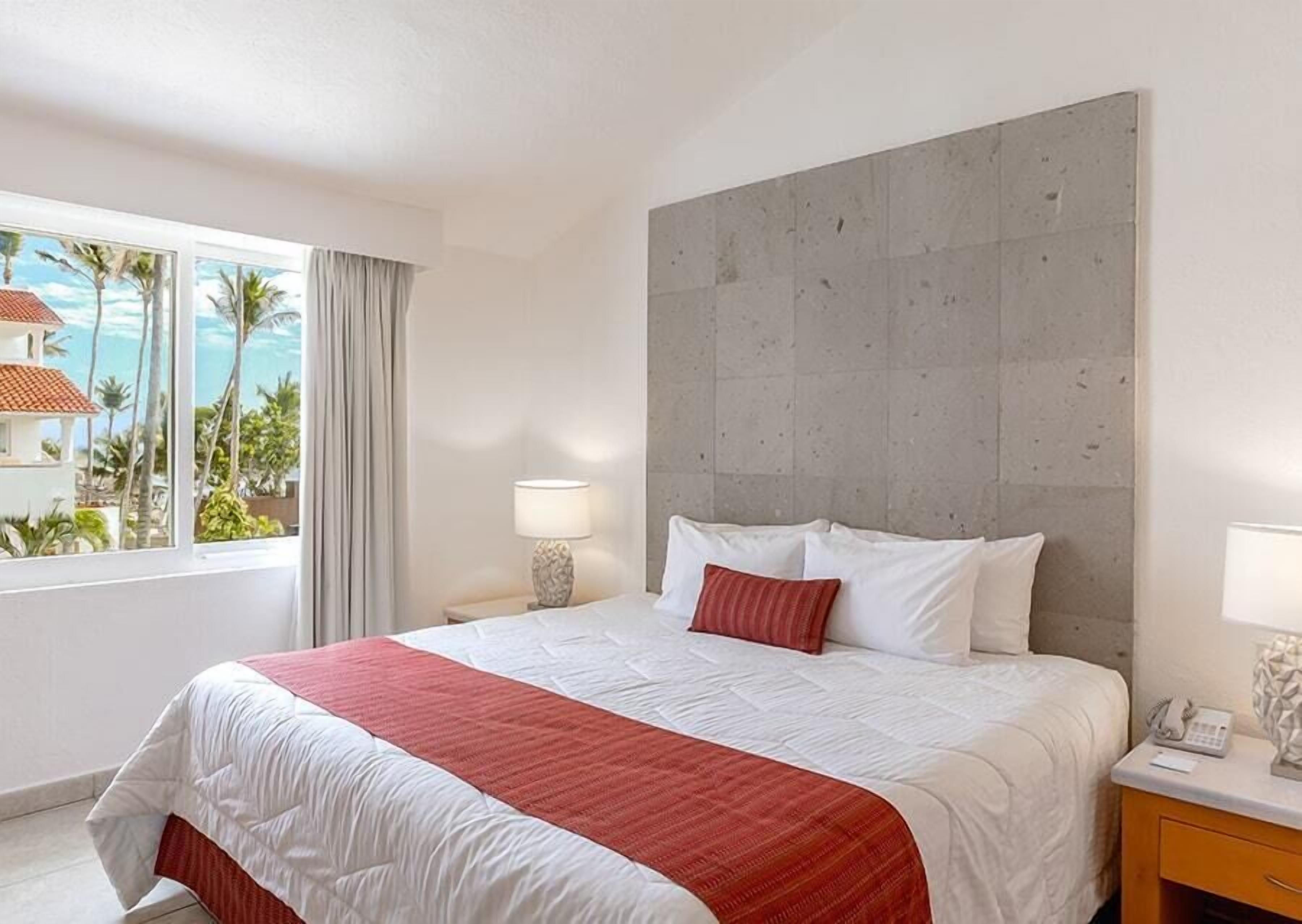 A hotel room with a large bed, a view of palm trees and buildings, and a grey headboard.