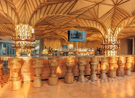 Zona de bar con techo alto y vigas de madera, que presenta una barra central con múltiples estantes de botellas y una pantalla de televisión en la parte superior.