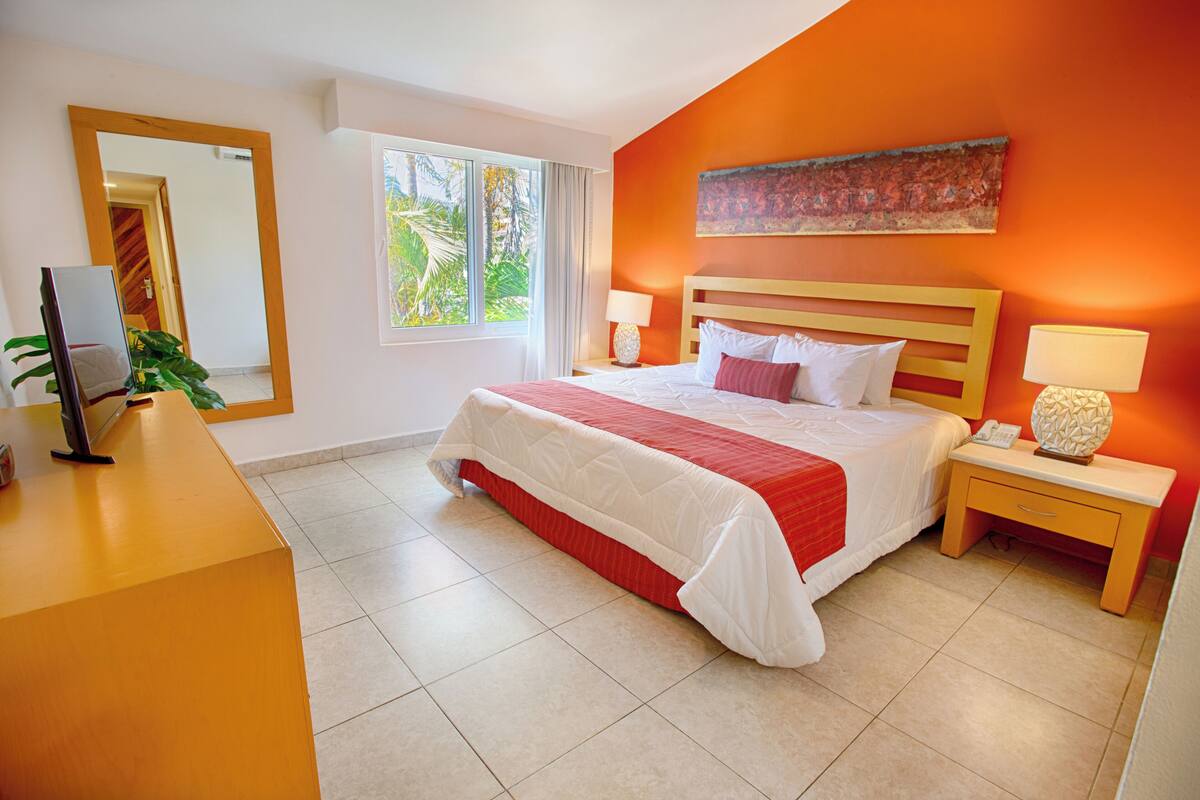A hotel room with a large bed, a wooden nightstand, a desk, and a window with a view of palm trees.