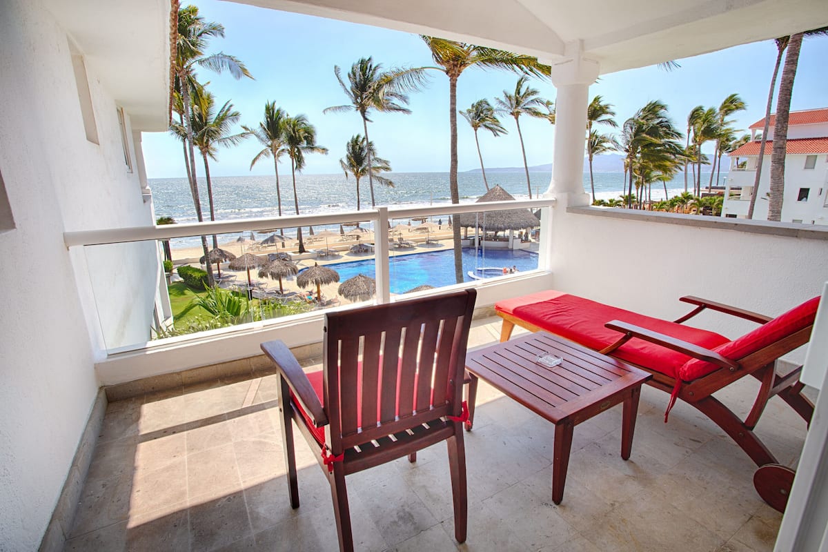 A balcony with a view of the beach and pool, wooden furniture, and a thatched-roof structure.