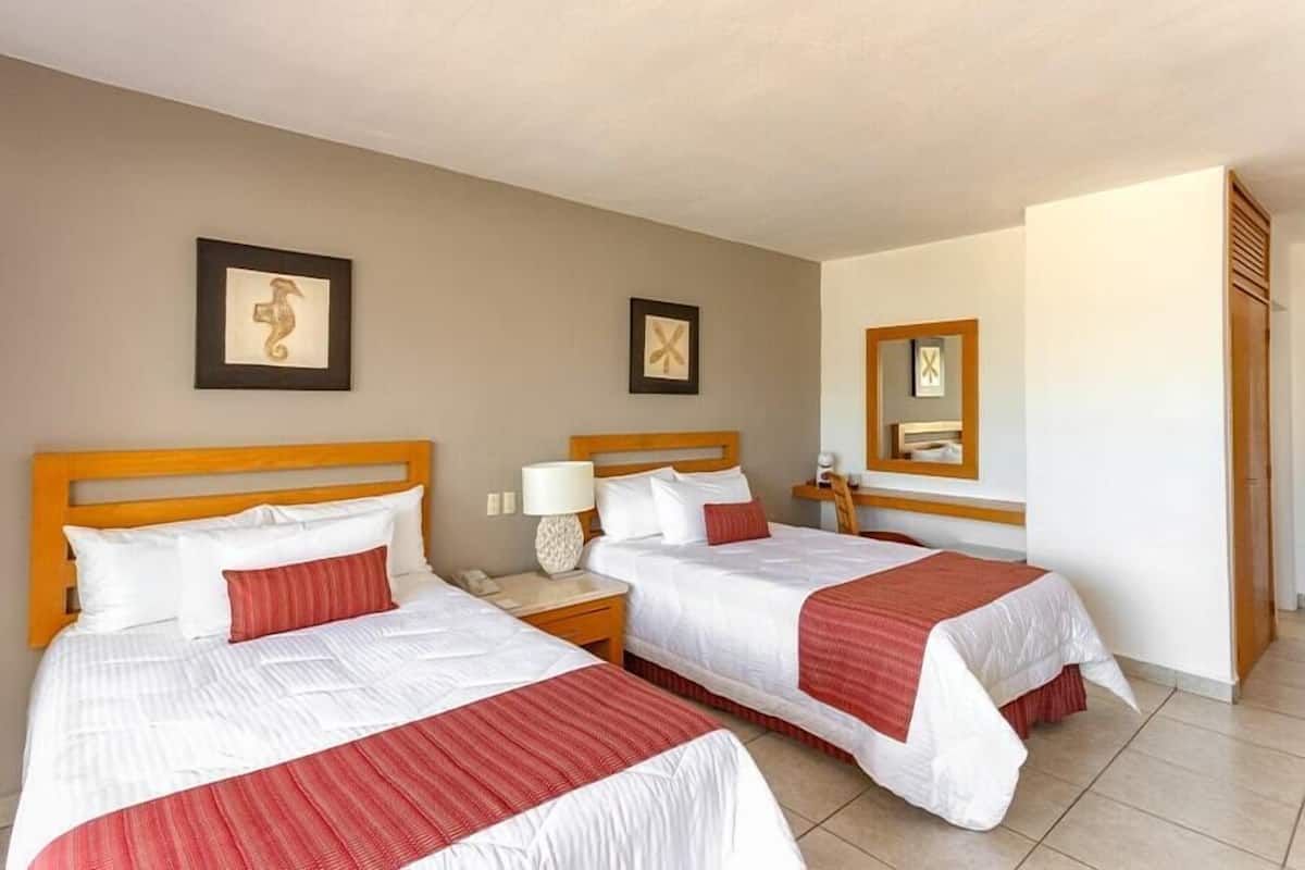 A hotel room with two beds, each with a wooden headboard, white bedding, and red accents. There are bedside tables with lamps, framed artwork on the walls, and a mirror above the bed.