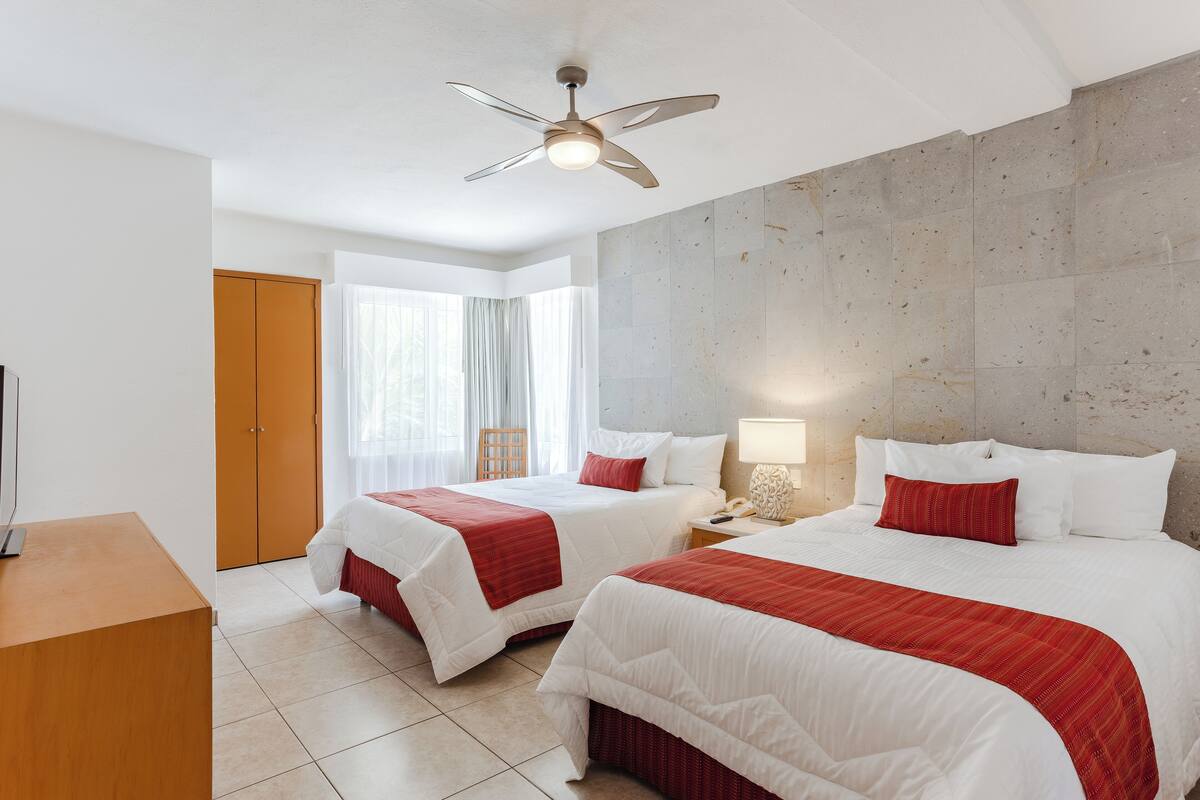 A hotel room with two beds, a ceiling fan, a wooden desk, and a view of the outside through large windows.