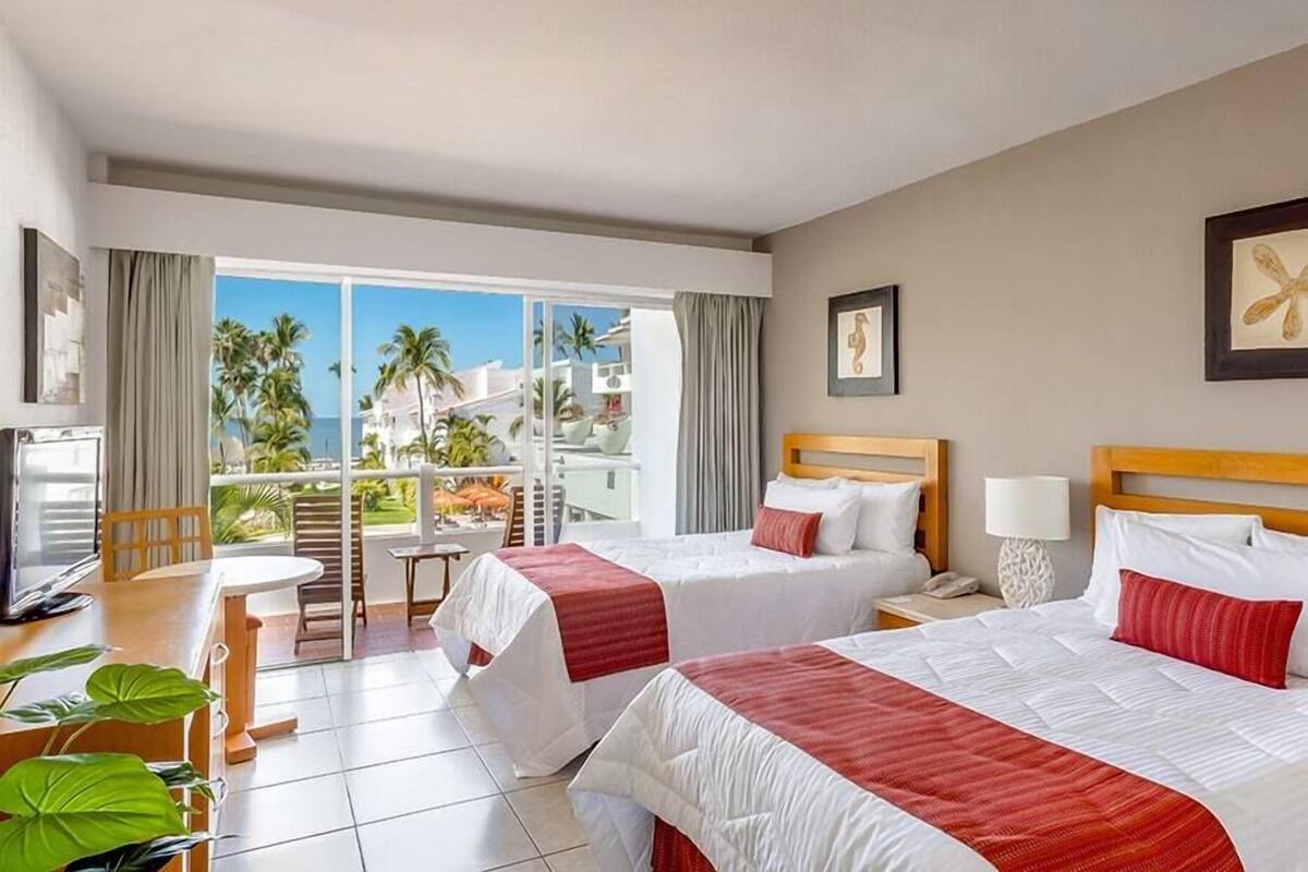 A hotel room with two beds, a desk, and a balcony with a view of palm trees and buildings.