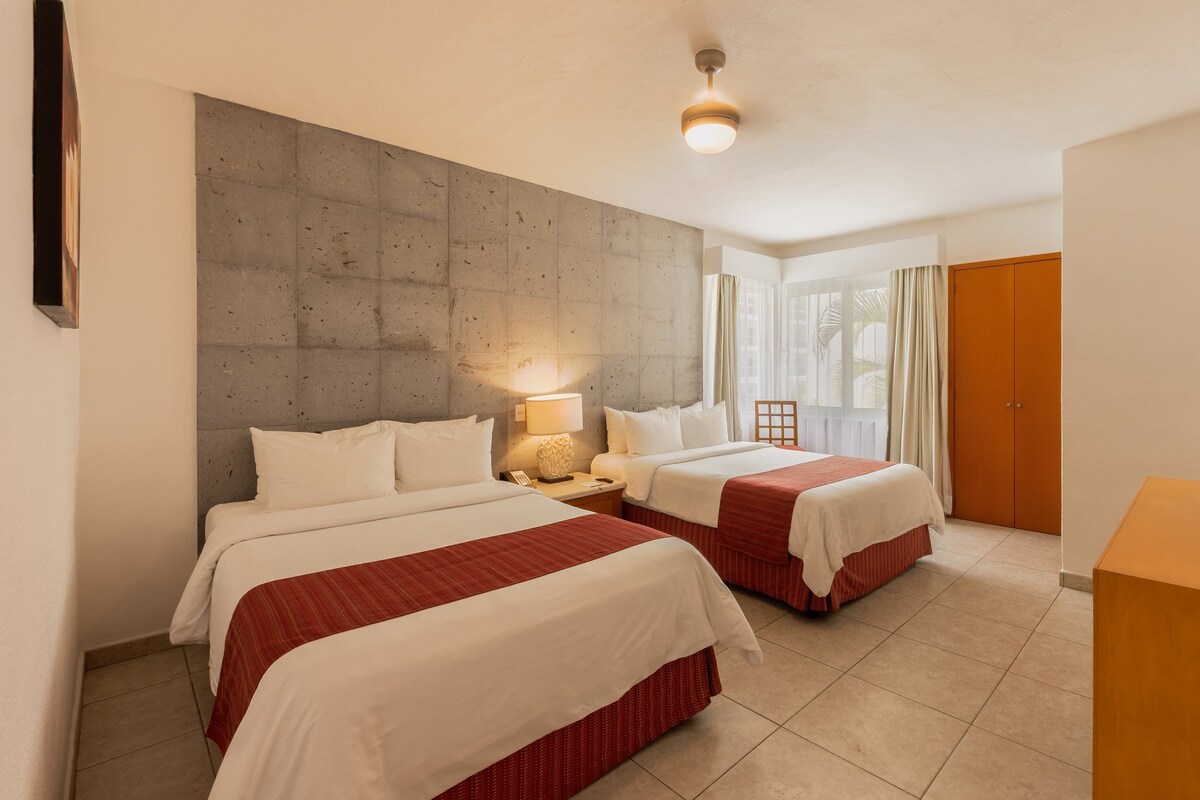 Habitación de hotel con dos camas, una pared de acento de concreto, ventilador de techo y ventana con cortinas.