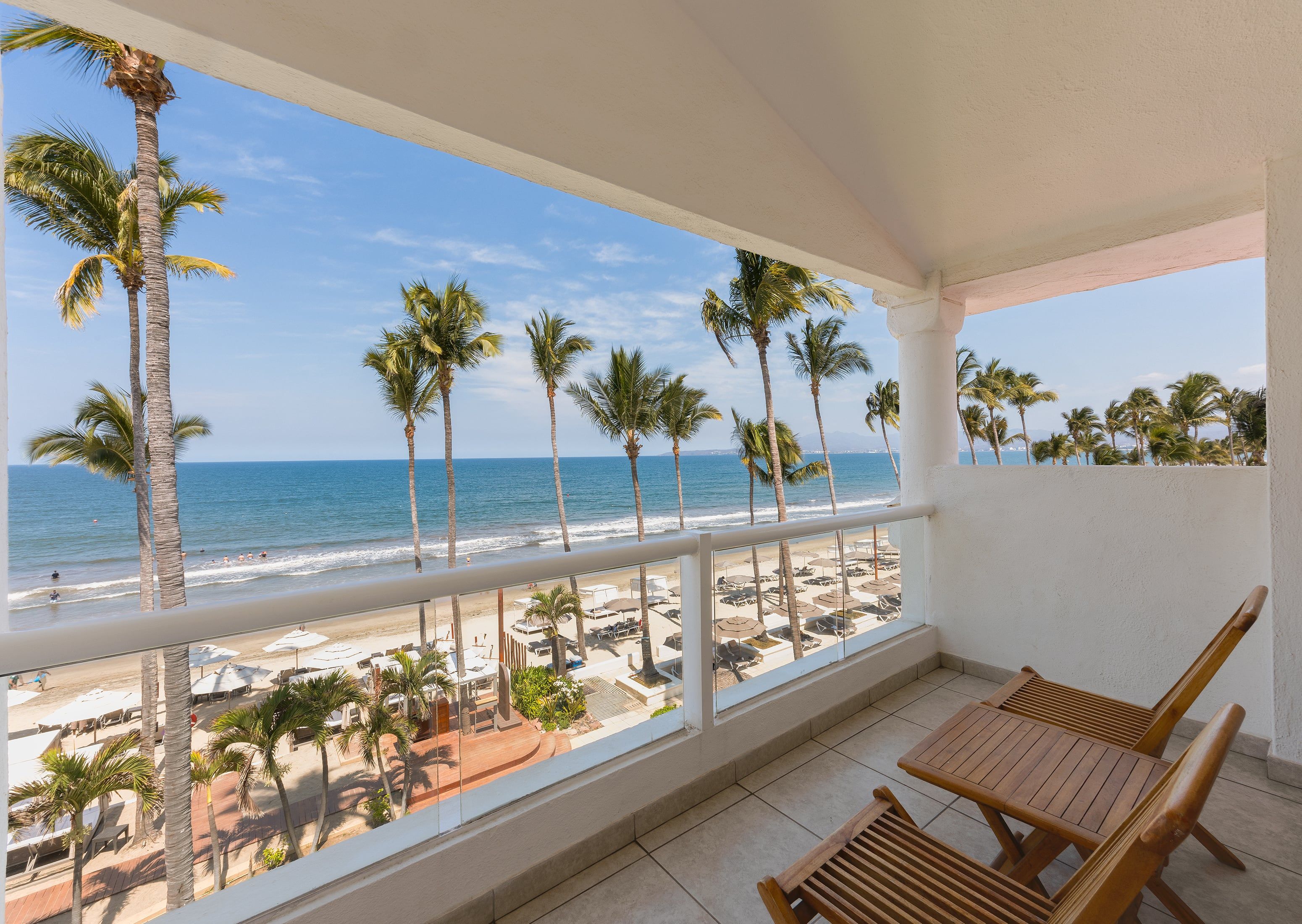 Ein Balkon mit Blick auf Strand und Meer, mit Palmen, Liegestühlen und Sonnenschirmen.