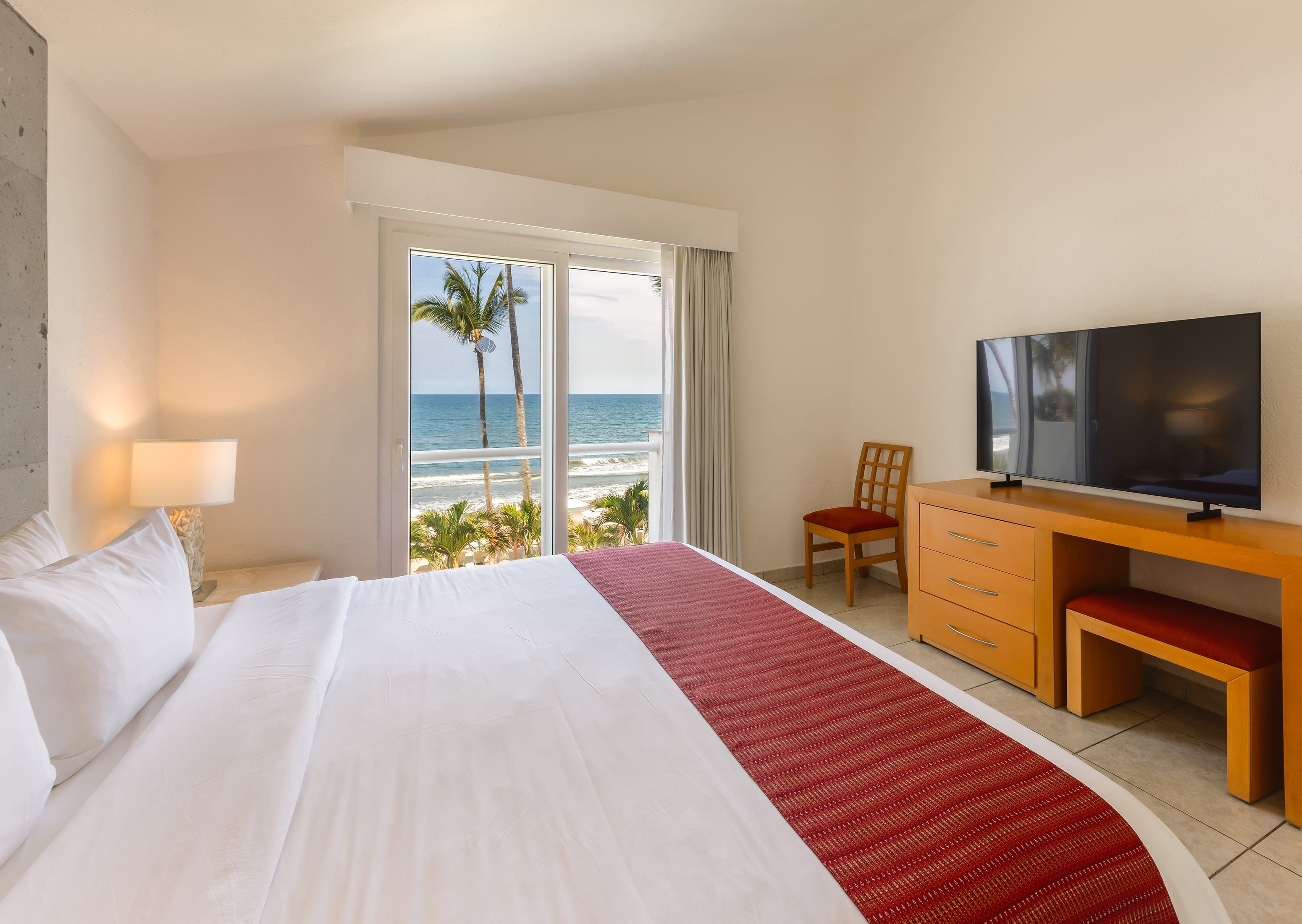 A hotel room with a bed, a desk, a chair, a television, and a view of the ocean.
