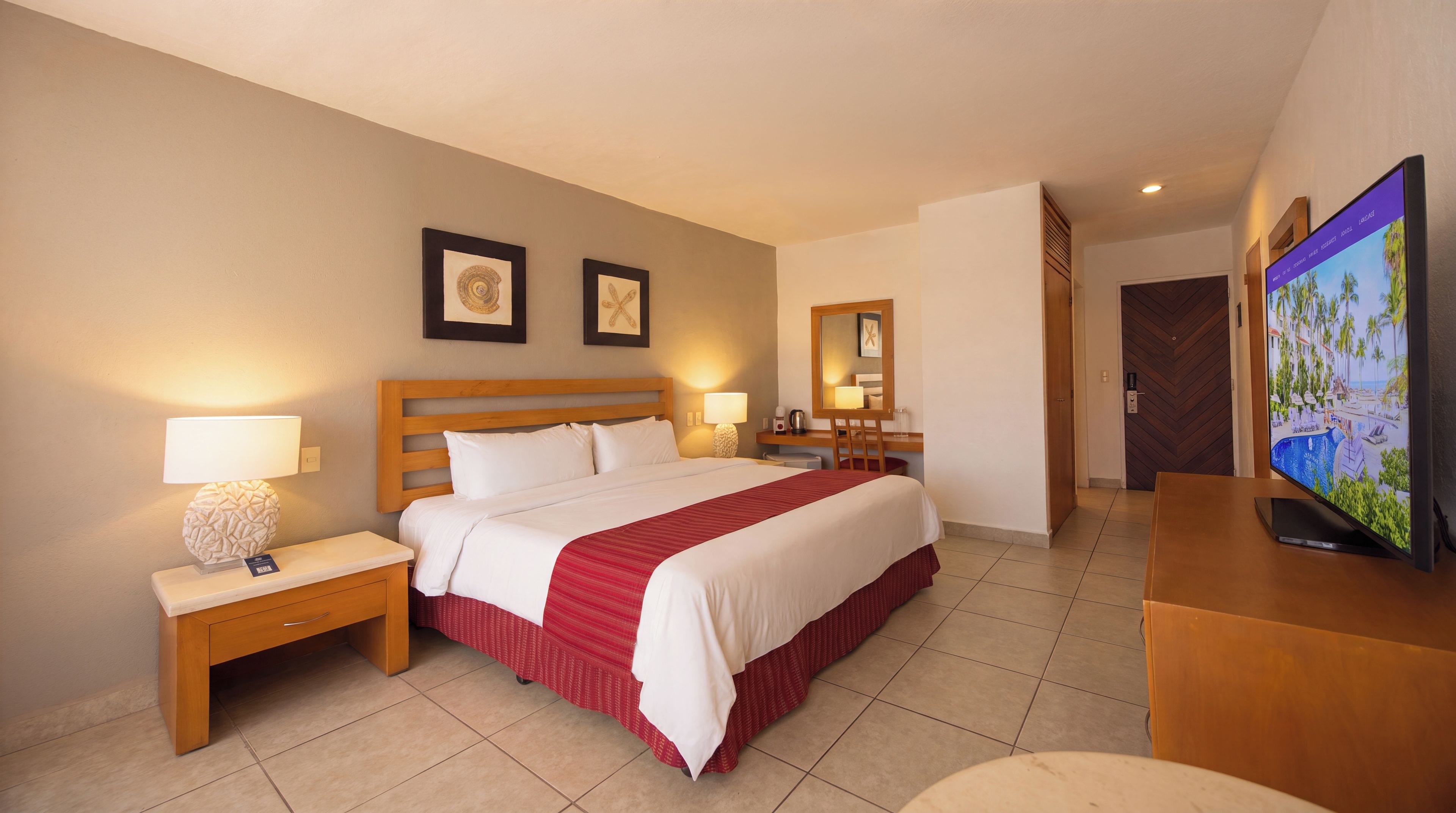 Una habitación de hotel con cama, mesitas de noche, escritorio, televisor y puerta de baño.