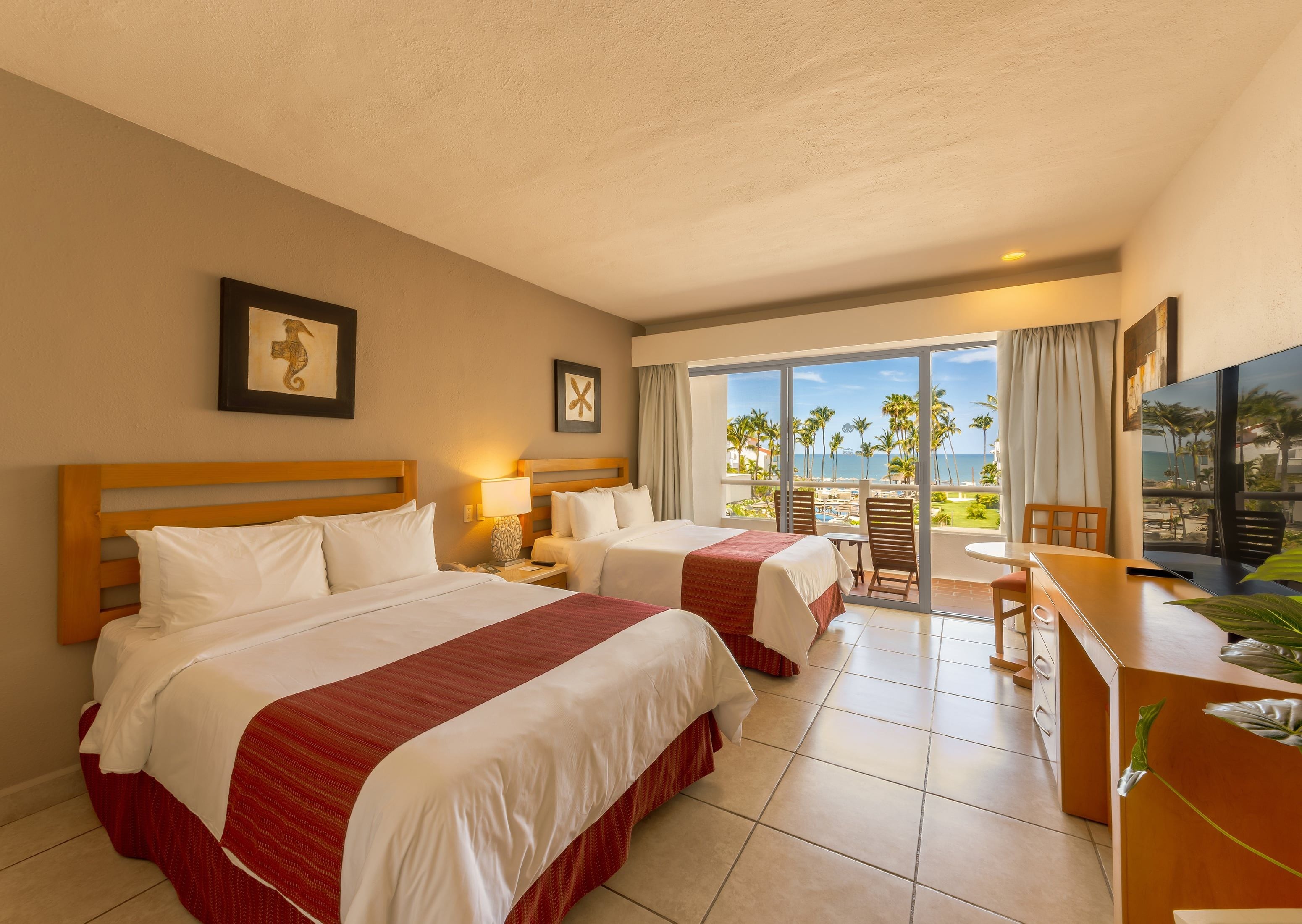 A hotel room with two beds, a desk, and a balcony with a view of the beach.