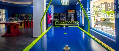 Una mesa de billar con una luz de neón encima, rodeada de máquinas de arcade y una ventana con vista al exterior.