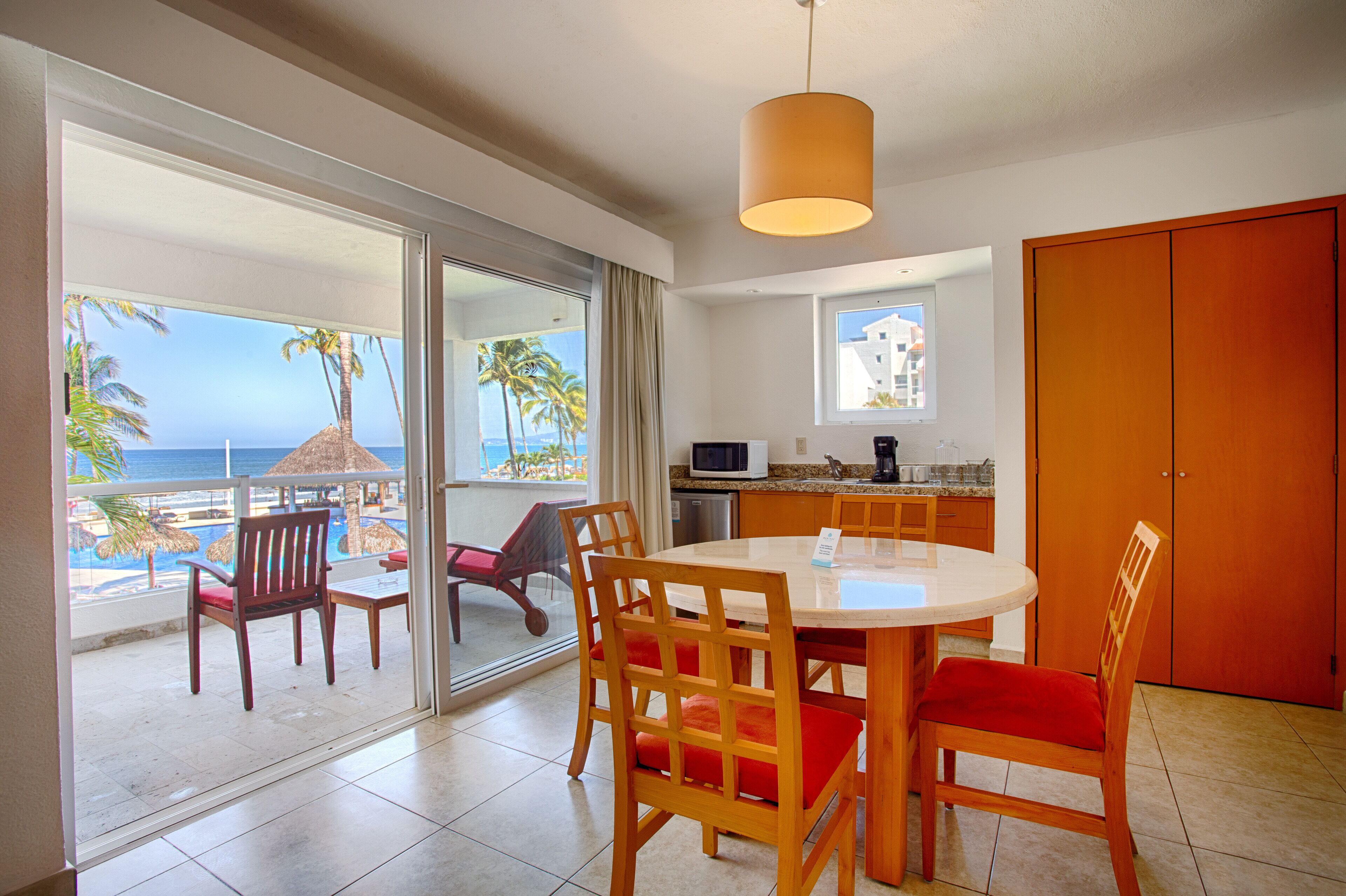 A room with a dining table, chairs, a kitchen area, and a view of the beach and ocean.