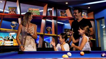 Une table de billard avec des billes, une famille qui joue et des étagères remplies de livres et de magazines.