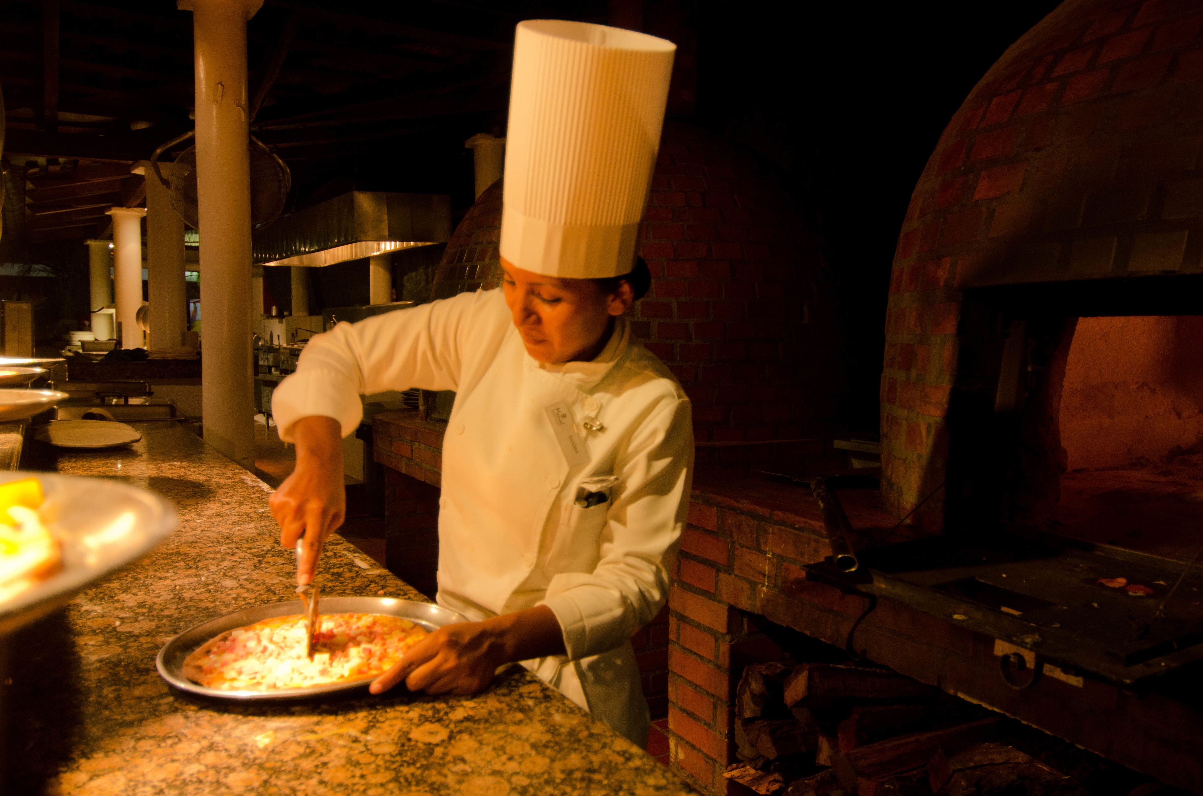 A chef in a white uniform with a tall hat, preparing food on a grill in a kitchen with brick walls and a large oven in the background.